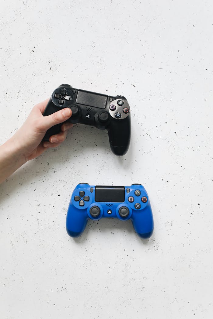 A hand holds a black PlayStation controller next to a blue one on a bright white surface.